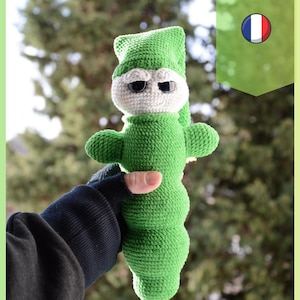 Puede incluir: Muñeca de ganchillo verde con cara blanca, ojos negros y gorro verde. La muñeca se sostiene en una mano con una manga negra. La imagen incluye el texto "Patron PDF" y un icono de la bandera francesa.