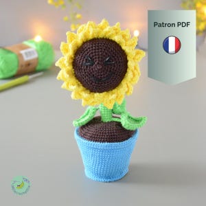 Op de afbeelding: Een gehaakte zonnebloem in een blauwe pot. De zonnebloem heeft een gele bloemblaadjeskroon, een bruin midden met een lachend gezicht en groene bladeren en stengel. De pot is lichtblauw. De afbeelding bevat ook een bol groen garen en een haaknaald.
