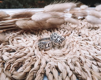May include: Two silver rings with floral designs, possibly crafted from repurposed silverware, are displayed on a woven mat. The rings showcase detailed flower patterns and are presented with dried ornamental grass, giving a rustic feel.