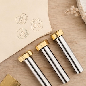 May include: Three cylindrical metal stamping tools with brass heads and silver shafts. Each tool features a unique engraved design on the face. A piece of light tan leather shows three distinct impressions: a heart with initials LP, a hexagon with initials CC, and a circular wreath with initials LR.