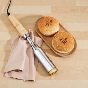 May include: A branding iron with a wooden handle and a stainless steel head, next to a pink cloth. Two sesame seed hamburger buns are on a wooden tray. One bun has "TOM'S BBQ" and a logo, the other has "A/K" and a logo.