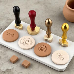 以下が含まれることがあります： 黒、赤、金色の木製ハンドルが付いたワックスシールスタンプのコレクション。スタンプは、さまざまなエンボス加工のデザインと、丸いワックスシールのイニシャルとともに表示されます。2つの小さなワックスキューブも存在します。