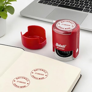May include: A red self-inking stamp with the text "NATIONALS 27 JUN 2026 CHASE WON!" is next to a red stamp holder and a notebook with stamped impressions. A laptop is in the background.