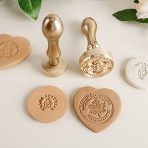 Puede incluir: Dos sellos metálicos dorados con mangos de madera y varias piezas de cuero estampadas. Un sello tiene un diseño de hoja de arce, y el otro tiene un diseño circular. Las piezas de cuero tienen varias formas, incluyendo un corazón y un círculo.