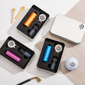 Peut inclure: Coffrets cadeaux sur le thème du golf dans des boîtes en métal argenté. Chaque ensemble comprend un objet cylindrique, une petite bouteille et un relève-pitch avec le logo "GOLFSTINA".