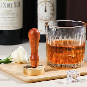 Op de afbeelding: Een houten en messing cocktailstamper staat op een houten plank naast een glas amberkleurige vloeistof met ijsblokjes. Een witte bloem en twee wijnflessen staan op de achtergrond. De stamper heeft een lichtbruine houten handgreep.