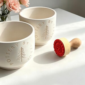 May include: Two cream-coloured ceramic bowls with the text "POTTERY BY TRUK" and a tree design. A wooden stamp with a red floral pattern is next to the bowls. Pink flowers in a glass vase are in the background.