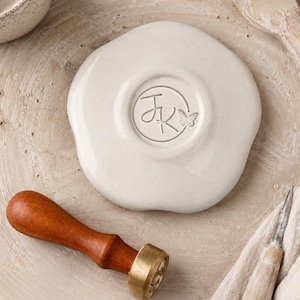 May include: A round, off-white ceramic piece with an irregular, organic edge. The center features a circular embossed design containing the letters JK and a small butterfly icon. A wooden handle wax seal stamp with a brass head is positioned nearby.