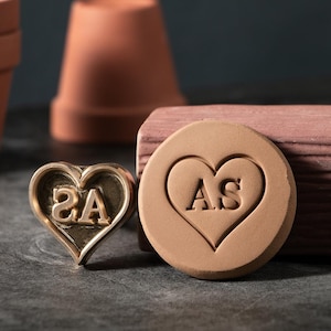 Könnte beinhalten: Ein herzförmiger Metallstempel mit den Buchstaben "SA" in einem Herzen, neben einem runden, hellbraunen Tonstempel mit den Buchstaben "AS" in einem Herzen. Der Hintergrund umfasst Terrakottatöpfe und einen Holzblock.