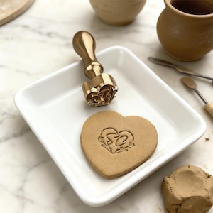 Könnte beinhalten: Ein herzförmiger Stempel aus Messing mit Holzgriff liegt auf einem weißen quadratischen Teller. Ein herzförmiger Keks mit den Initialen "SC" und floralem Design befindet sich neben dem Stempel. Töpferwerkzeuge und Ton sind im Hintergrund.