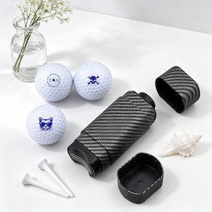 Könnte beinhalten: Weiße Golfbälle mit blauen Grafiken, darunter eine Katze mit Sonnenbrille, ein Totenkopf mit gekreuzten Knochen und Initialen. Ein schwarz-grauer Golfballhalter und weiße Golf-Tees sind ebenfalls vorhanden.