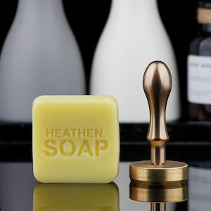 May include: A square, pale yellow bar of soap with the word "HEATEN SOAP" embossed on its surface. A gold-colored stamp with a rounded handle and circular base is positioned next to the soap. The background features blurred white and gray bottles.