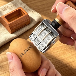 Könnte beinhalten: Ein braunes Ei mit dem Stempel "30 JAN 2026" wird neben einem Datumsstempel mit Holzgriff gehalten. Im Hintergrund befindet sich eine Holzkiste mit dem Text "PAR AVION". Der Datumsstempel ist silber und schwarz.