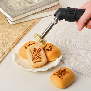 Puede incluir: Un hierro de marcar con cabeza de latón se utiliza para imprimir diseños en bollos dorados. Los bollos están dispuestos en un plato blanco y presentan varios diseños, incluyendo un tenedor y una espátula cruzados, y el nombre "Vincent".