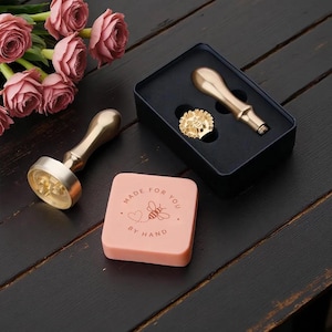 Könnte beinhalten: Ein Wachssiegel-Set mit zwei goldfarbenen Metallgriffen und einem runden Messingstempel mit Bienenmotiv. Ein quadratischer, hellrosa Wachsblock ist mit einer Bienen-Grafik, einem Herz und dem Text "MADE FOR YOU BY HAND" in Dunkelrosa bedruckt.