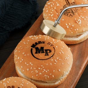 Könnte beinhalten: Nahaufnahme von Sesam-Hamburgerbrötchen mit dem Text "MADE BY Mp" auf dem oberen Brötchen. Ein Metall-Branding-Werkzeug ist über den Brötchen positioniert, um das obere Brötchen zu brandmarken. Die Brötchen liegen auf einem Holzschneidebrett.