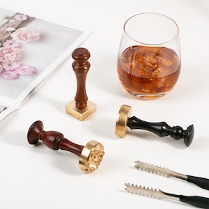 Op de afbeelding: Een assortiment zegelstempels met houten handvatten en messing stempelkoppen. Een glas amberkleurige vloeistof met ijsblokjes en een ijstang zijn ook aanwezig. De items zijn gerangschikt op een wit oppervlak.