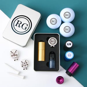Puede incluir: Una lata plateada con las iniciales "RG" en un círculo, un juego de marcadores de bola de golf con diferentes iniciales, un recuperador de bola de golf, una pequeña botella de líquido, un tubo dorado y tres bolas de golf con las iniciales "RG" impresas en ellas.