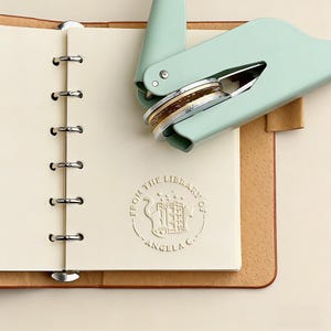 Puede incluir: Una herramienta de encuadernación verde menta con una pila de monedas de oro y plata, colocada sobre un cuaderno abierto. El cuaderno tiene una cubierta de color claro con un sello de biblioteca en relieve y el texto "FROM THE LIBRARY OF...ANGELA C."