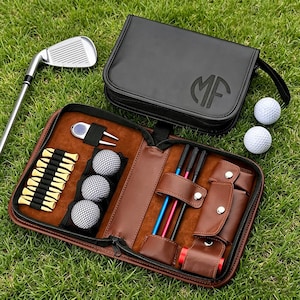 Könnte beinhalten: Ein schwarzes Golf-Zubehöretui mit Reißverschluss und eingeprägten Buchstaben MF auf der Vorderseite, das drei weiße Golfbälle, eine Pitchgabel aus Metall, zehn gelbe Golftees und drei farbige Ausrichtungsstäbe sowie einen silbernen Golfschläger enthält.