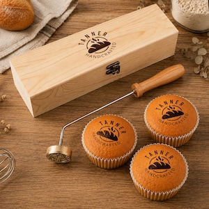 May include: A metal branding iron with a wooden handle, a rectangular wooden storage box, and two cupcakes. The branding iron and the top of the wooden box feature a circular logo with a loaf of bread and the text TANNER HANDCRAFTED.