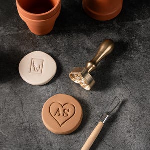 May include: A collection of pottery tools and materials, including a heart-shaped stamp with "AS" initials, a stamp with a square design, and a brass handle stamp. Two small terracotta pots are also visible.