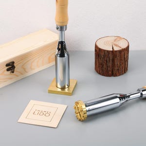 May include: A branding iron with a wooden handle and a brass stamp head. The iron is silver and gold in color. A wooden box and a small wooden coaster with the text "THAMES LIDO" are also in the image. A small wooden log is also in the image.