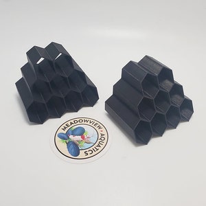 May include: Two black, honeycomb-shaped structures, possibly for aquariums, sit next to a round sticker with the text "Meadowview Aquatics" and a fish illustration. The structures are made of a dark material and have a geometric design.