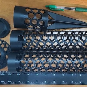 May include: A set of black plastic components featuring a hexagonal honeycomb pattern, including two cylindrical tubes, a pointed ground stake, a circular base, and a cap, displayed alongside a green pen and a ruler measuring up to 12 inches.
