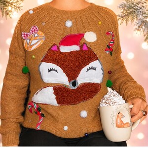Puede incluir: Un suéter de punto color canela con una gran aplicación central de un zorro con un gorro de Papá Noel rojo. El suéter está decorado con bastones de caramelo de tela, un adorno con lazo y pequeños pompones blancos. Se sostiene una taza color crema.