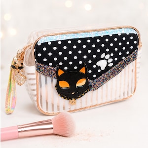 Op de afbeelding: Een rechthoekige transparante toilettas met witte strepen, voorzien van een zwart paneel met witte stippen en een zwarte kattenkop-applicatie met oranje ogen. De tas heeft een glitterende rand, een pootafdruk-detail en een polsbandje.