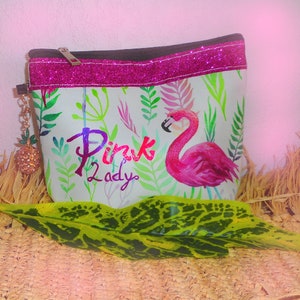 May include: A white cosmetic bag featuring a pink flamingo design and the text "Pink Lady." The bag has a black zip, a pink glitter trim, and a gold pineapple charm. The background includes green and pink foliage.