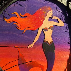 May include: A purple, orange and black mermaid with long red hair and a black tail with orange and purple accents. The mermaid is painted on a wooden oval.