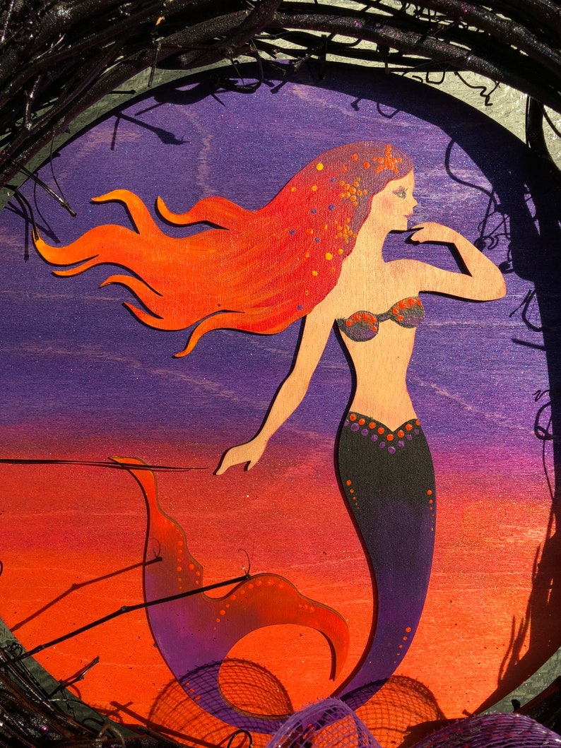 May include: A purple, orange and black mermaid with long red hair and a black tail with orange and purple accents. The mermaid is painted on a wooden oval.