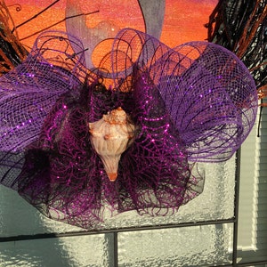 May include: A purple and black glittery mesh wreath with a seashell in the centre. The wreath is decorated with black and brown twigs.