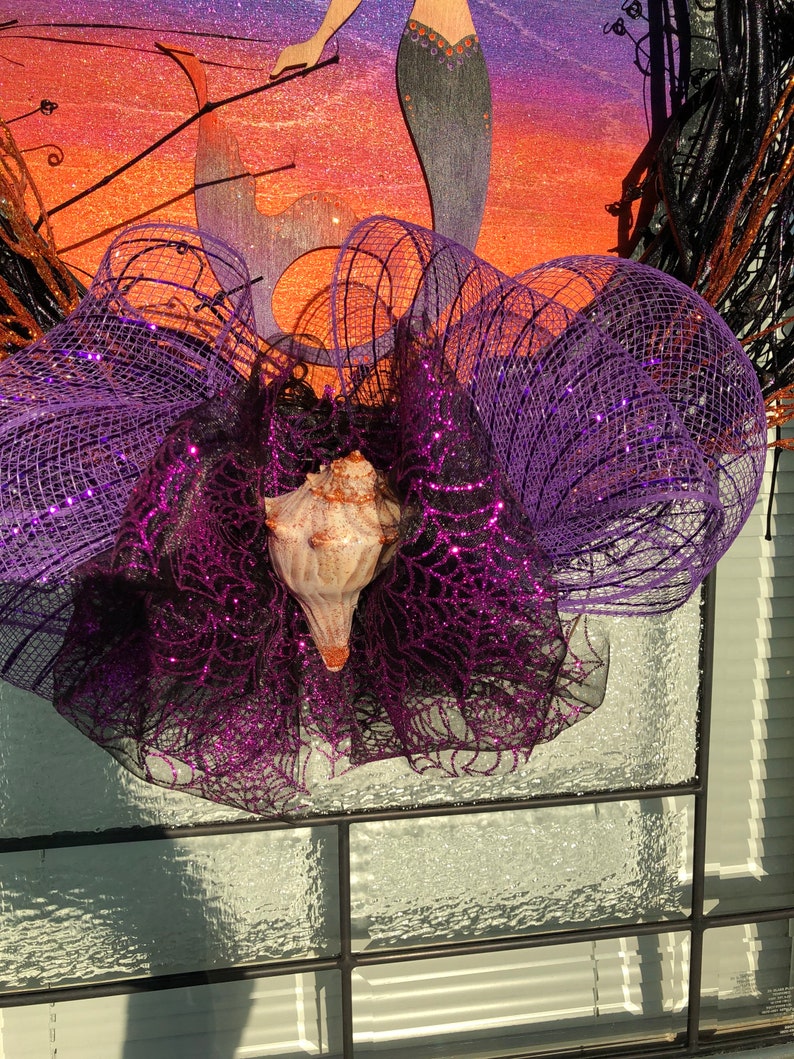 May include: A purple and black glittery mesh wreath with a seashell in the centre. The wreath is decorated with black and brown twigs.