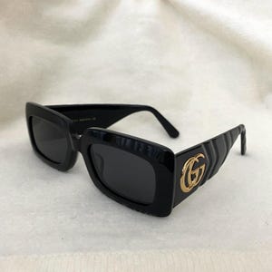 May include: Black rectangular sunglasses with dark lenses. The frames are thick and glossy, with a gold-colored double G logo on the temples. The sunglasses are set against a white background.