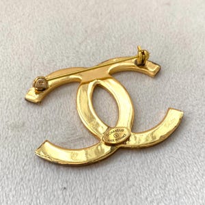 Pode incluir: Um broche de metal dourado em forma de log&oacute;tipo CC entrela&ccedil;ado. A parte de tr&aacute;s apresenta um mecanismo de alfinete horizontal e uma placa oval gravada com o texto CHANEL, um s&iacute;mbolo de copyright e as palavras MADE IN FRANCE.