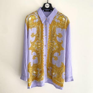 Puede incluir: Camisa de manga larga de color lila claro con un estampado barroco dorado y botones dorados. La etiqueta Versace es visible en la percha. La camisa está hecha de un material sedoso.