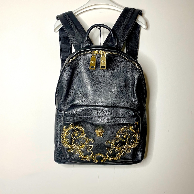 May include: Black leather backpack with gold-tone hardware. The front pocket features a decorative gold stud and emblem design. The backpack has a top handle, adjustable shoulder straps, and a zippered main compartment.