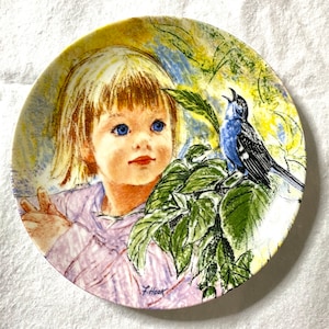 May include: A vintage ceramic plate featuring a colourful illustration of a young girl with blonde hair and blue eyes, a blue bird, and green leaves. The plate is signed "F. Hook" in the bottom right corner.