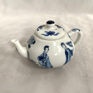May include: A small, white ceramic teapot with blue floral and figure designs. The teapot has a curved spout, a small handle, and a lid with a dark blue knob. The design features Asian-inspired figures and floral patterns.
