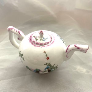 May include: A white porcelain teapot with a round body and a floral design. The teapot has a pink lid with a small hole in the top. The teapot is decorated with pink, blue, and green flowers.