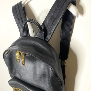 May include: Black leather backpack with gold-tone hardware, including zippers and buckles. The shoulder straps feature a textured design. The backpack is hanging on a white hanger.