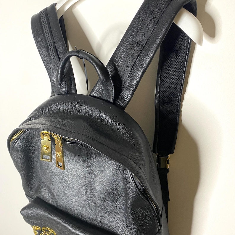 May include: Black leather backpack with gold-tone hardware, including zippers and buckles. The shoulder straps feature a textured design. The backpack is hanging on a white hanger.