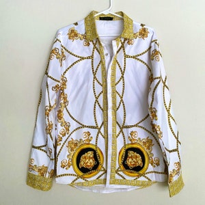 Puede incluir: Camisa blanca con estampado barroco dorado y negro. La camisa presenta un diseño repetido de cadenas y flores, con dos medallones en la parte inferior. El cuello y los puños tienen un patrón de greca dorada.