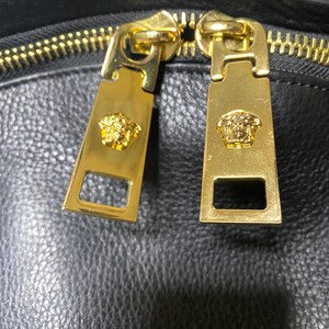 May include: Close-up of a black leather bag with gold-colored zippers. The zipper pulls are rectangular with a gold Medusa head emblem. The bag's leather has a textured appearance.