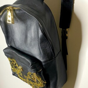 May include: Black leather backpack with gold-tone hardware. Features a front pocket adorned with gold embellishments and a gold zipper. The backpack has a top handle and adjustable shoulder straps. The zippers and hardware have a gold finish.