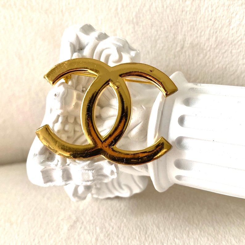 Pode incluir: Um broche de metal em tom dourado com o ic&oacute;nico design do log&oacute;tipo de CC entrela&ccedil;ados. A superf&iacute;cie polida e refletora do emblema cria uma silhueta suave e curva. O broche est&aacute; posicionado sobre uma base de pilar decorativa branca.