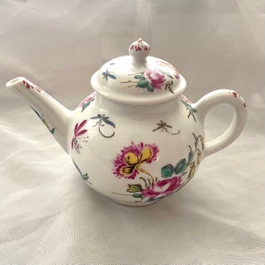 May include: A white porcelain teapot with a floral design. The teapot has a round body with a lid and a spout. The floral design features pink, red, yellow, and green flowers and leaves. There are also small dragonfly designs on the teapot.